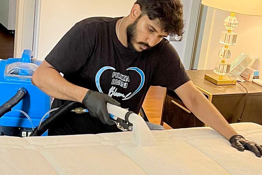 Uomo che pulisce un materasso con un pulitore a vapore in una camera da letto, indossando guanti e una maglietta nera - igienizzazione professionale materasso non sfoderabile Milano per una pulizia e una freschezza profonde.