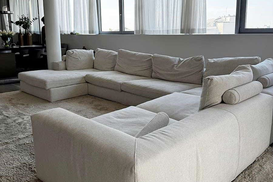 Grande divano componibile beige in un soggiorno moderno con finestre alte e tende trasparenti. Approfitta dello sconto pulizia divani Milano per mantenere il tuo divano impeccabile e accogliente.