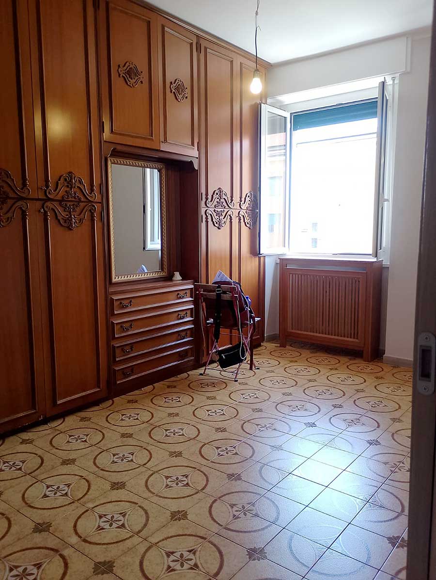 Camera con armadi in legno decorati, pavimento in piastrelle decorate, finestra aperta e una sedia con borse vicino al termosifone - ideale per sgombero e pulizia post imbiancatura Milano.