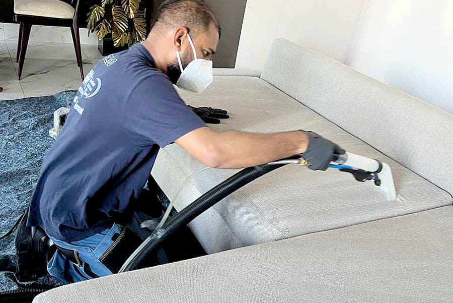 Un uomo con guanti e mascherina pulisce un divano chiaro con un detergente professionale per tappezzeria, offrendo Pulizia Divani a domicilio a Milano.