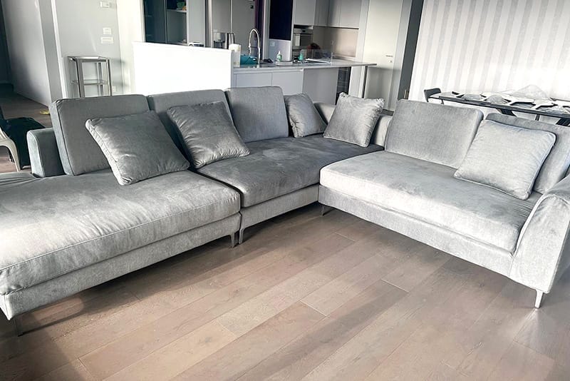 Un grande divano componibile grigio con diversi cuscini in un soggiorno moderno con pavimento in legno, perfetto per case a Milano vicino a domicilio Amendola Fiera e ideale per comodi servizi di pulizia divano.