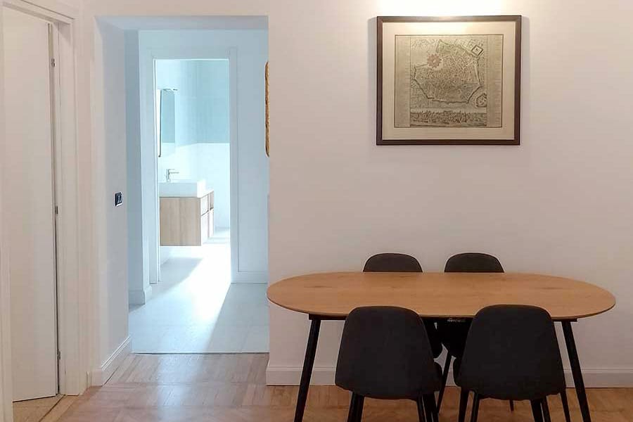 Un tavolo da pranzo in legno con quattro sedie nere si trova vicino a una parete bianca con opere d'arte incorniciate, accanto a un luminoso corridoio: l'ideale per chi cerca pulizia appartamenti arredati Milano e uno spazio abitativo elegante e pulito.