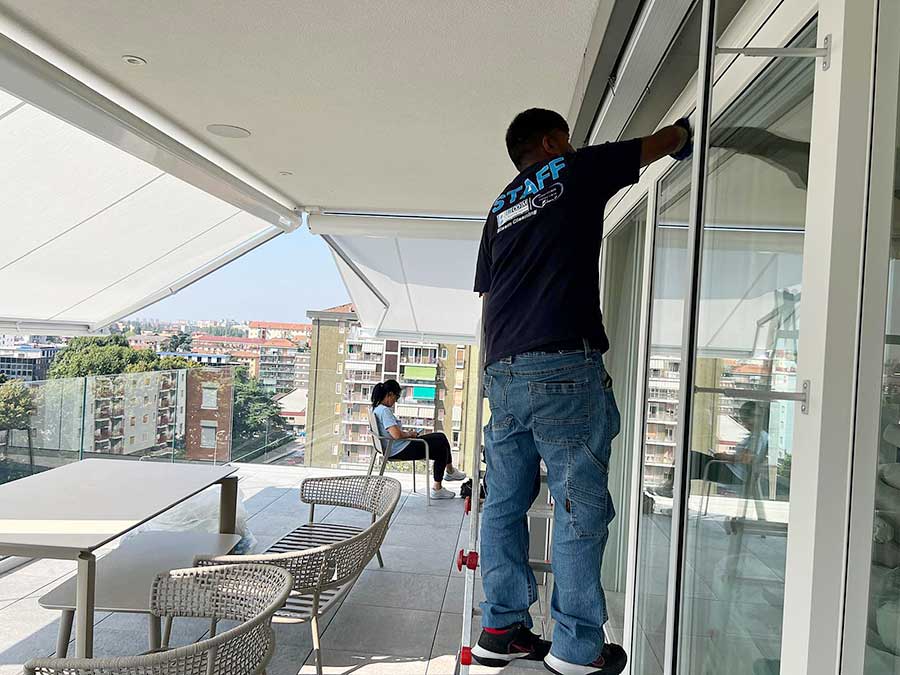 Un uomo pulisce le finestre di un balcone in Zona Moscova, a Milano, mentre una donna siede su una sedia all'esterno, con vista sui palazzi della città -ulizia appartamenti al suo meglio.