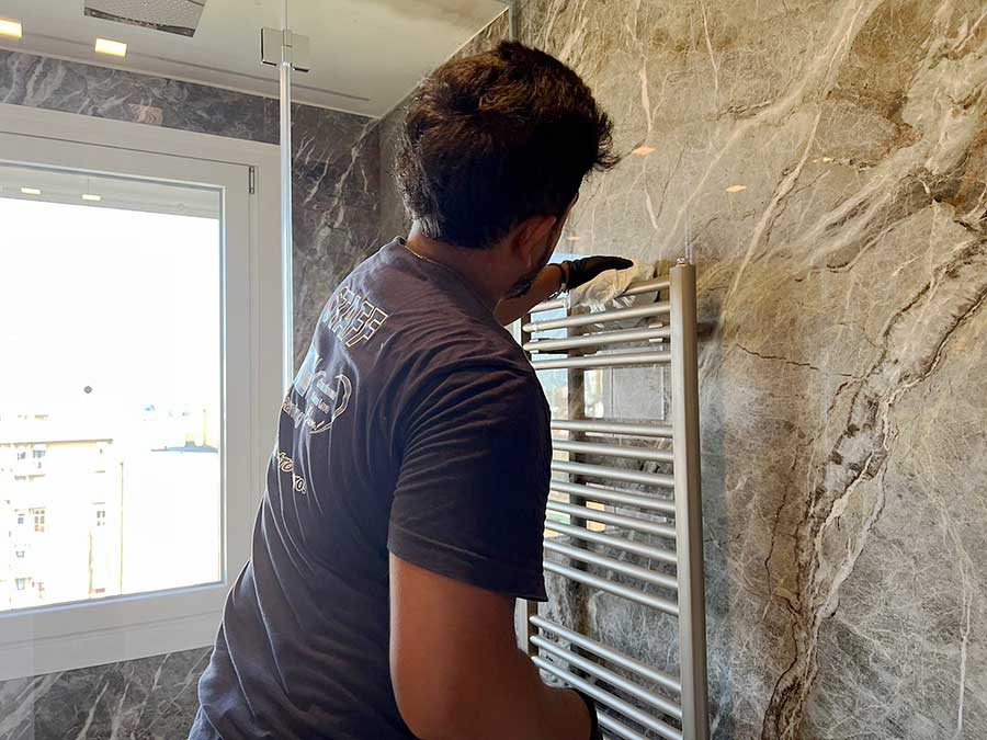 Una persona installa un portasciugamani metallico su una parete del bagno in marmo vicino a una finestra in un elegante appartamento di Milano in zona Moscova, dimostrando attenzione ai dettagli e agli standard di pulizia appartamenti.