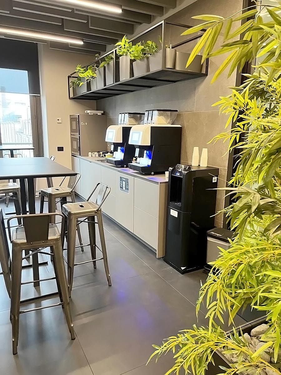 Una moderna sala riunioni in zona Solari, a Milano, con sedie e tavoli in metallo, macchine da caffè su un bancone, un distributore d'acqua, piante in vaso in alto e verde in primo piano. Le grandi finestre riempiono di luce naturale questo spazio ispirato alla pulizia degli uffici.