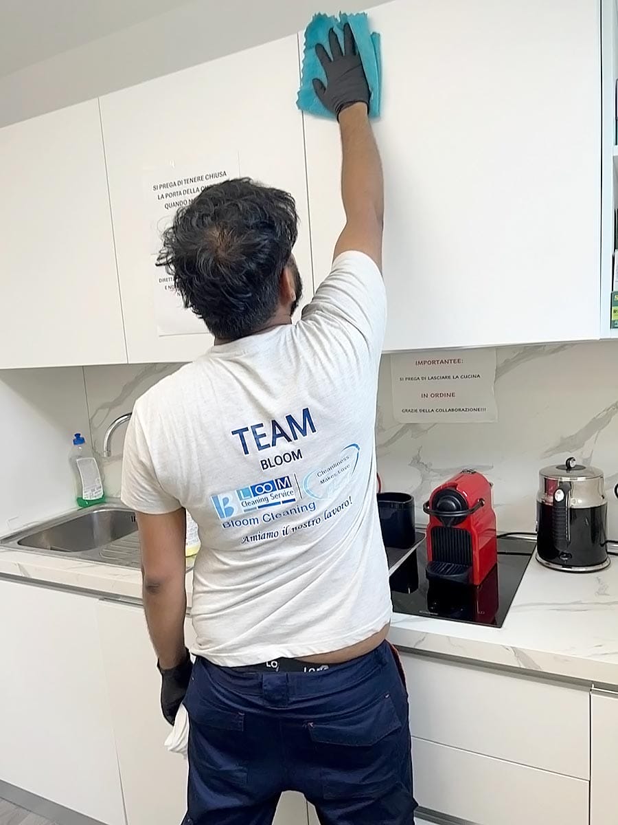 Una persona con guanti e maglietta "TEAM", di un'impresa pulizie uffici zona Isola Milano, pulisce i pensili superiori della cucina con un panno blu. Sul piano di lavoro sono appoggiati una macchina del caffè rossa e dei prodotti per la pulizia.