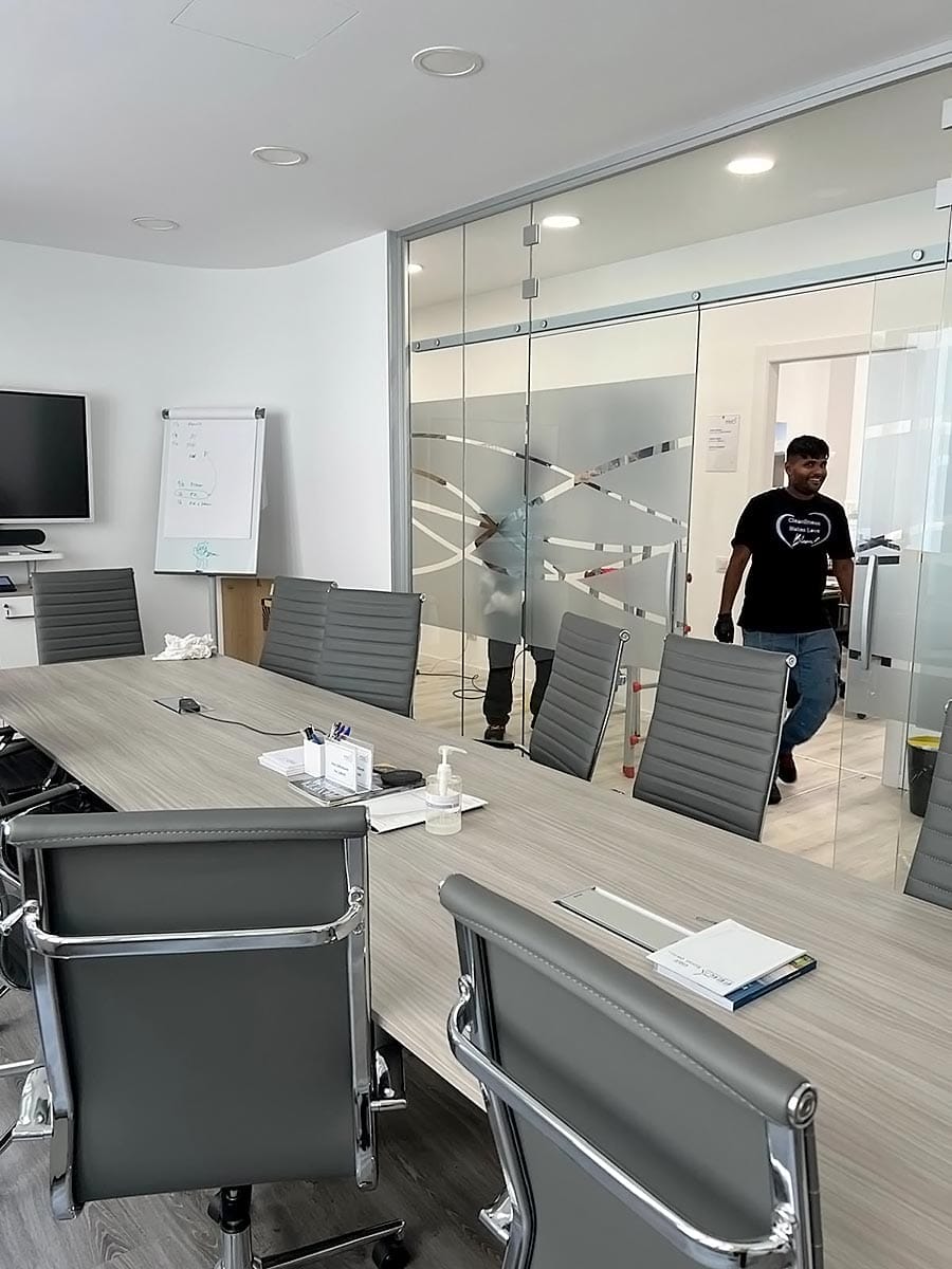 Una moderna sala conferenze con un lungo tavolo, sedie grigie e materiale d'ufficio, tenuta in ordine da un'impresa pulizie uffici zona Isola Milano. Due persone si vedono attraverso le pareti di vetro, con una lavagna e un televisore montati a parete.