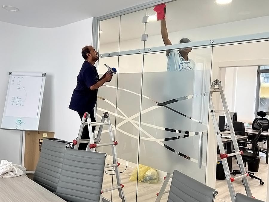 Due persone di un'impresa pulizie uffici zona Isola Milano puliscono una parete dell'ufficio in vetro con motivi geometrici, utilizzando una spatola e un panno rosso. Nello spazio di lavoro moderno si vedono una sedia da ufficio, un tavolo da riunione e una lavagna a fogli mobili.
