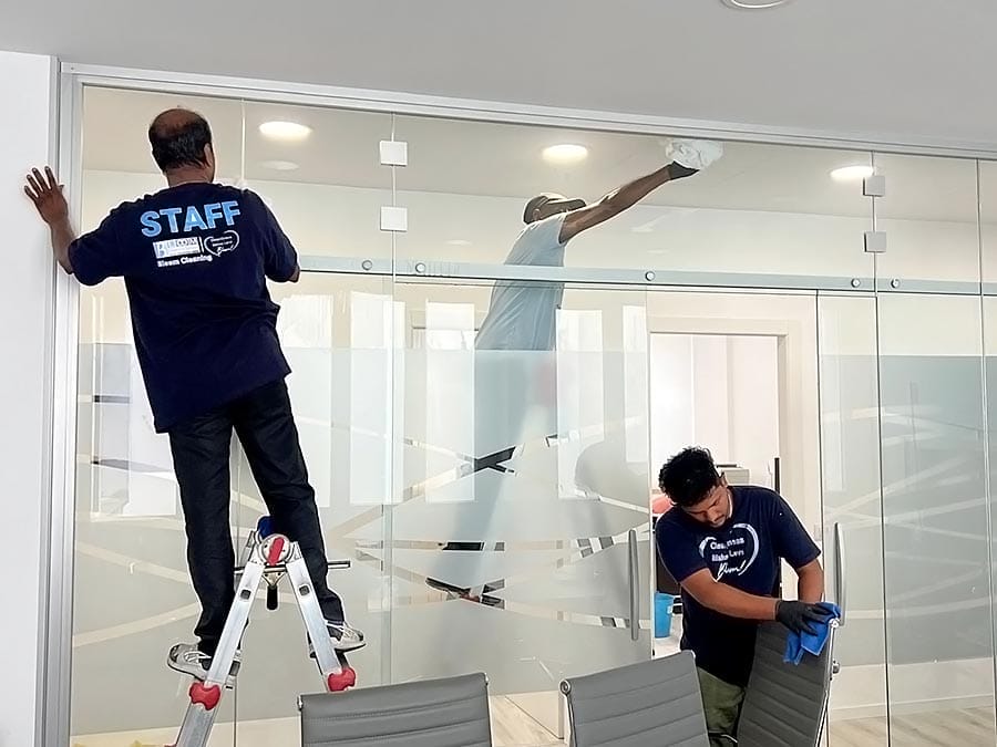 Tre addetti alle pulizie in camicia blu puliscono le pareti di vetro di un ufficio. Uno è in piedi su una scala a pioli, un altro pulisce in alto e il terzo pulisce i pannelli inferiori vicino alle sedie di un'impresa pulizie uffici zona Isola Milano.