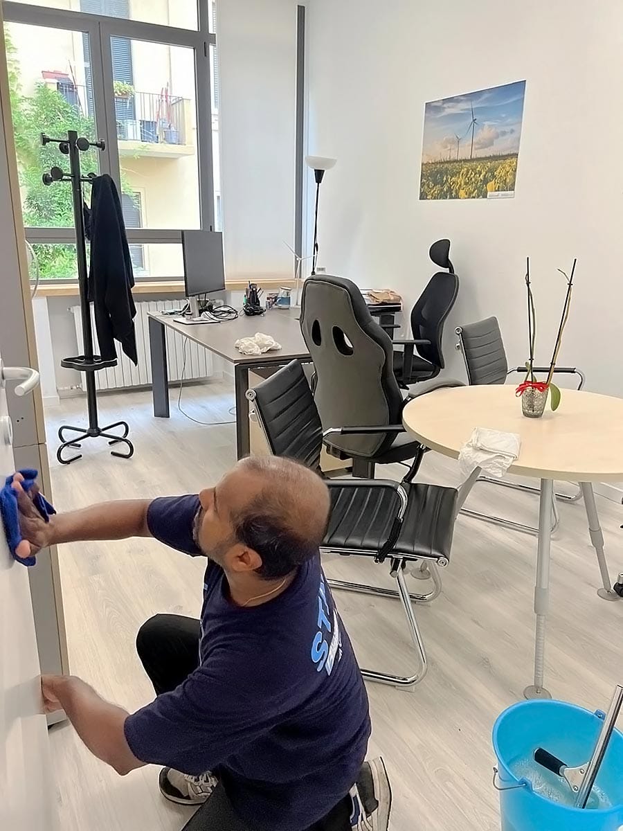 Una persona in camicia blu pulisce una porta in un ufficio moderno, mostrando la professionalità di un'impresa pulizie uffici viale Zara Milano. Scrivanie, sedie e luce naturale completano l'ordinato spazio di lavoro.