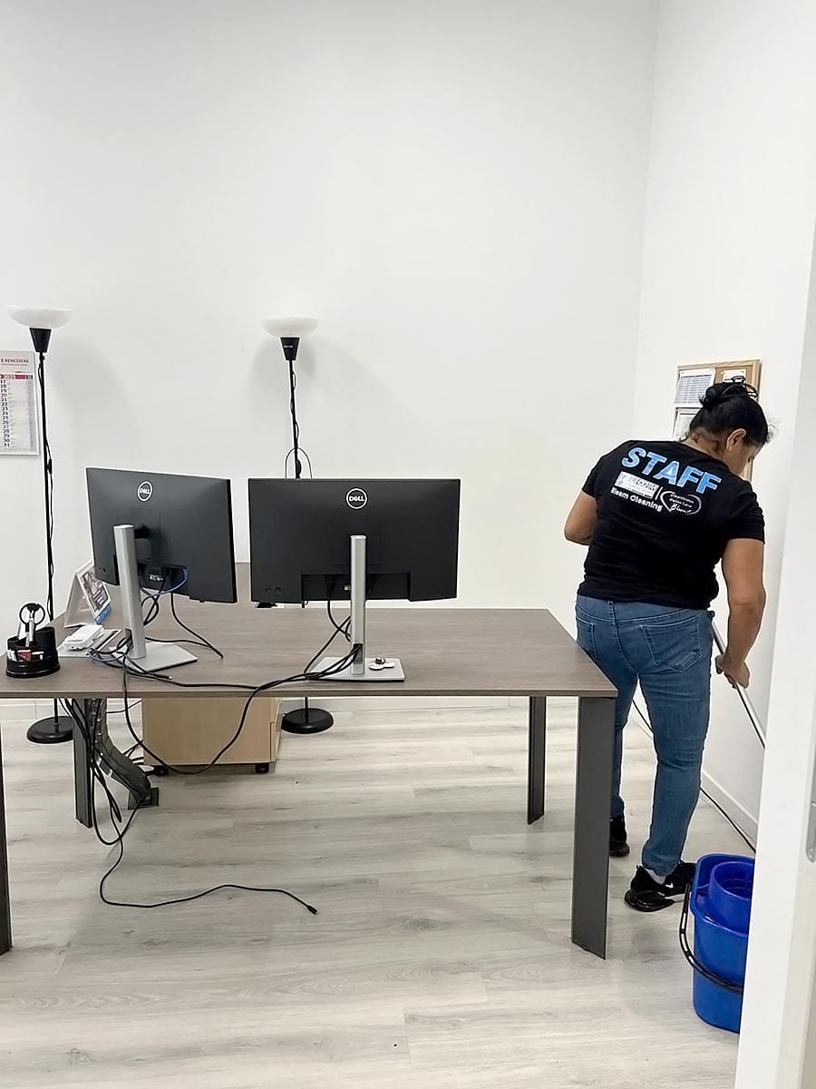 Una donna in maglietta "STAFF" pulisce il pavimento di un luminoso ufficio vicino a viale Zara Milano, con due monitor su una scrivania, un secchio blu e lampade a stelo, che mostrano la qualità del lavoro di un'impresa pulizie uffici viale Zara Milano.