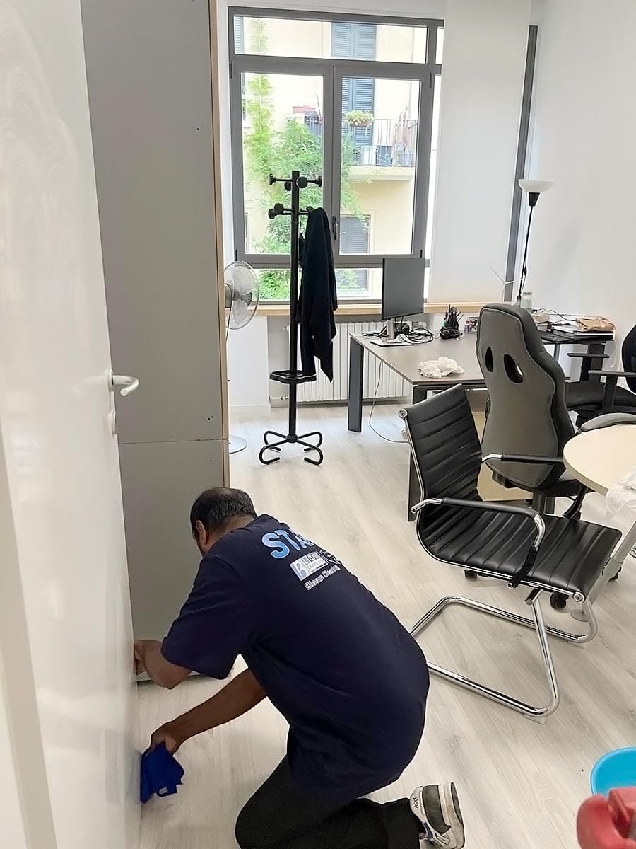 Una persona con una camicia blu navy è inginocchiata sul pavimento e pulisce vicino a una parete bianca in un ufficio moderno con sedie, scrivanie e un appendiabiti che mette in mostra la professionalità dell'impresa pulizie uffici zona Isola Milano.