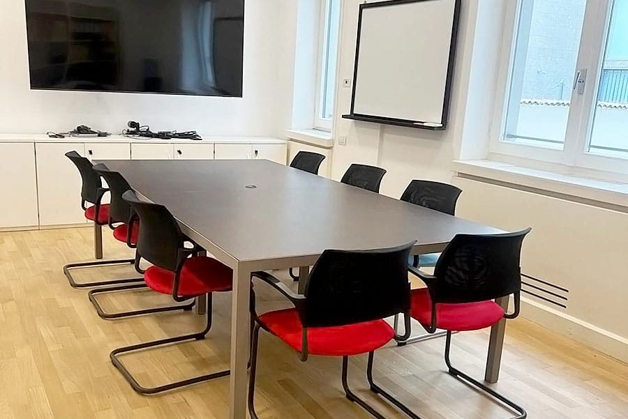 Una sala conferenze con un tavolo rettangolare, otto sedie nere con sedili rossi, un grande schermo, una lavagna e finestre illuminate dal sole, mantenuta pulita con pulizia uffici Milano e servizio pulizia programmato per uno spazio di lavoro accogliente.