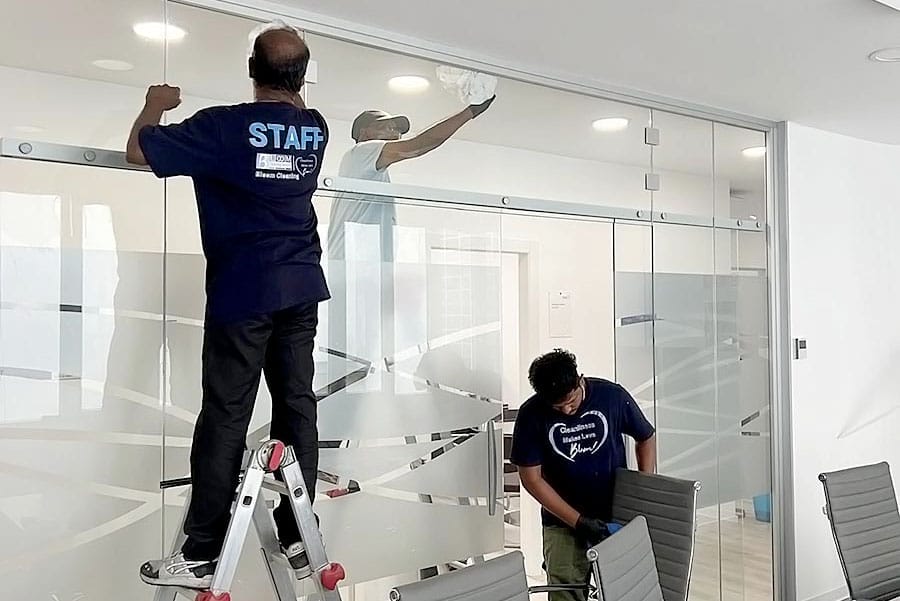 Tre addetti alle pulizie di un'impresa pulizie uffici zona Isola Milano stanno pulendo le grandi pareti di vetro di un ufficio moderno. Uno pulisce il vetro su una scaletta, un altro pulisce in piedi e un terzo pulisce un tavolo vicino alle sedie dell'ufficio.