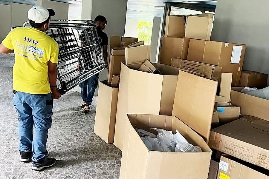 Due persone spostano grandi scatole di cartone e una rastrelliera metallica all'esterno di un edificio a Milano; una indossa una maglietta gialla "STAFF". Scatole di varie dimensioni sparse sul pavimento in pietra suggeriscono un servizio chiavi in mano per sgombero appartamento Milano.