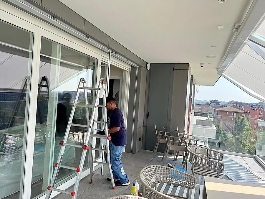 Una persona in piedi su un balcone accanto a una scala, sembra lavorare vicino a grandi porte di vetro. Sul pavimento sono presenti i prodotti per la pulizia di Pulizia Appartamenti, mentre sullo sfondo sedie e un tavolo si affacciano su edifici e alberi.