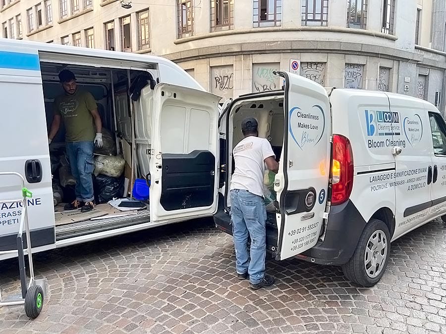 Due operai scaricano materiale di pulizia da due furgoni bianchi parcheggiati su una strada acciottolata di fronte a un edificio. I furgoni espongono i loghi "Bloom Cleaning", evidenziando i servizi di Pulizia Appartamenti e Sanificazione Divani.