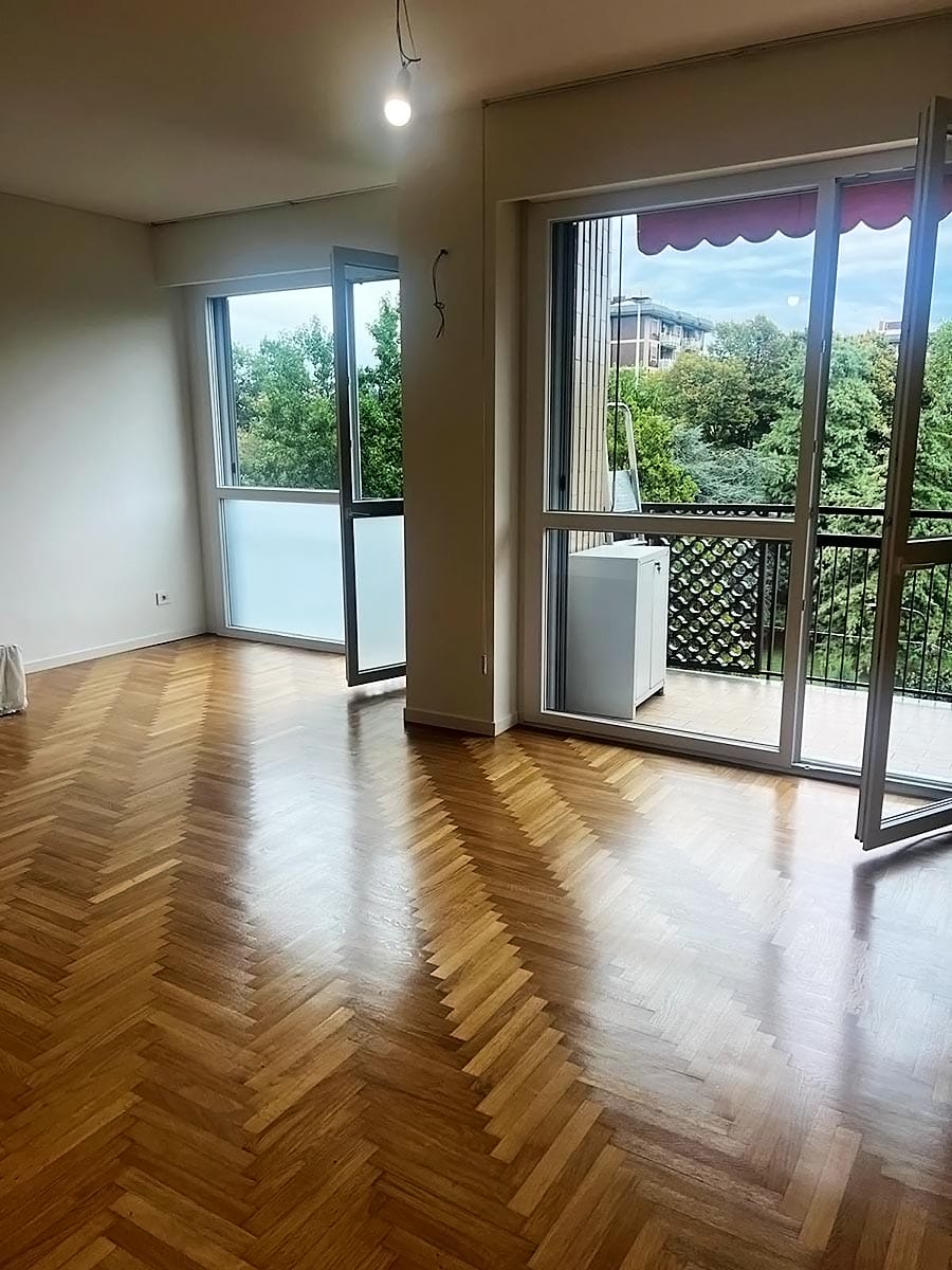 Locale luminoso e vuoto, con pavimento in legno a spina di pesce, ampie finestre e porte in vetro che si aprono su un balcone verdeggiante; due plafoniere sono visibili e una lavatrice si trova vicino al balcone: perfetto per Pulizia Appartamenti o Sanificazione Divani.