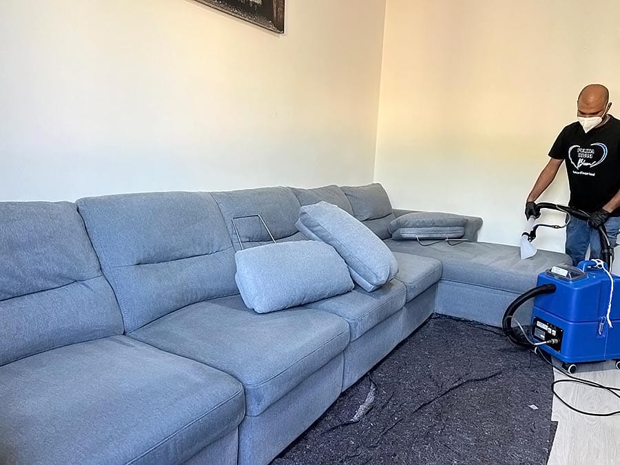 Una persona che indossa una maschera esegue la sanificazione divani, pulendo un grande divano componibile azzurro con una macchina professionale in una stanza luminosa. Alcuni cuscini vengono spostati e un tappetino scuro viene steso sul pavimento.