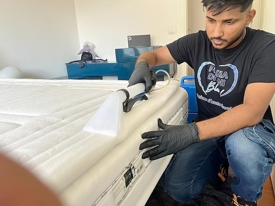 Un uomo che indossa guanti neri utilizza un dispositivo di pulizia per pulire a fondo un materasso bianco in una stanza luminosa. Sullo sfondo, una cassettiera blu e vari oggetti fanno pensare a Pulizia Appartamenti o Sanificazione Divani in corso.