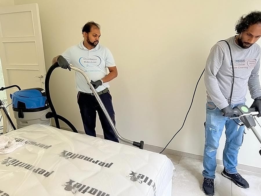 Due uomini in abiti da lavoro casual usano l'aspirapolvere per pulire a fondo un materasso in una stanza luminosa. La plastica ricopre parte del materasso e nelle vicinanze sono visibili vari materiali di pulizia per un lavoro efficiente e professionale.
