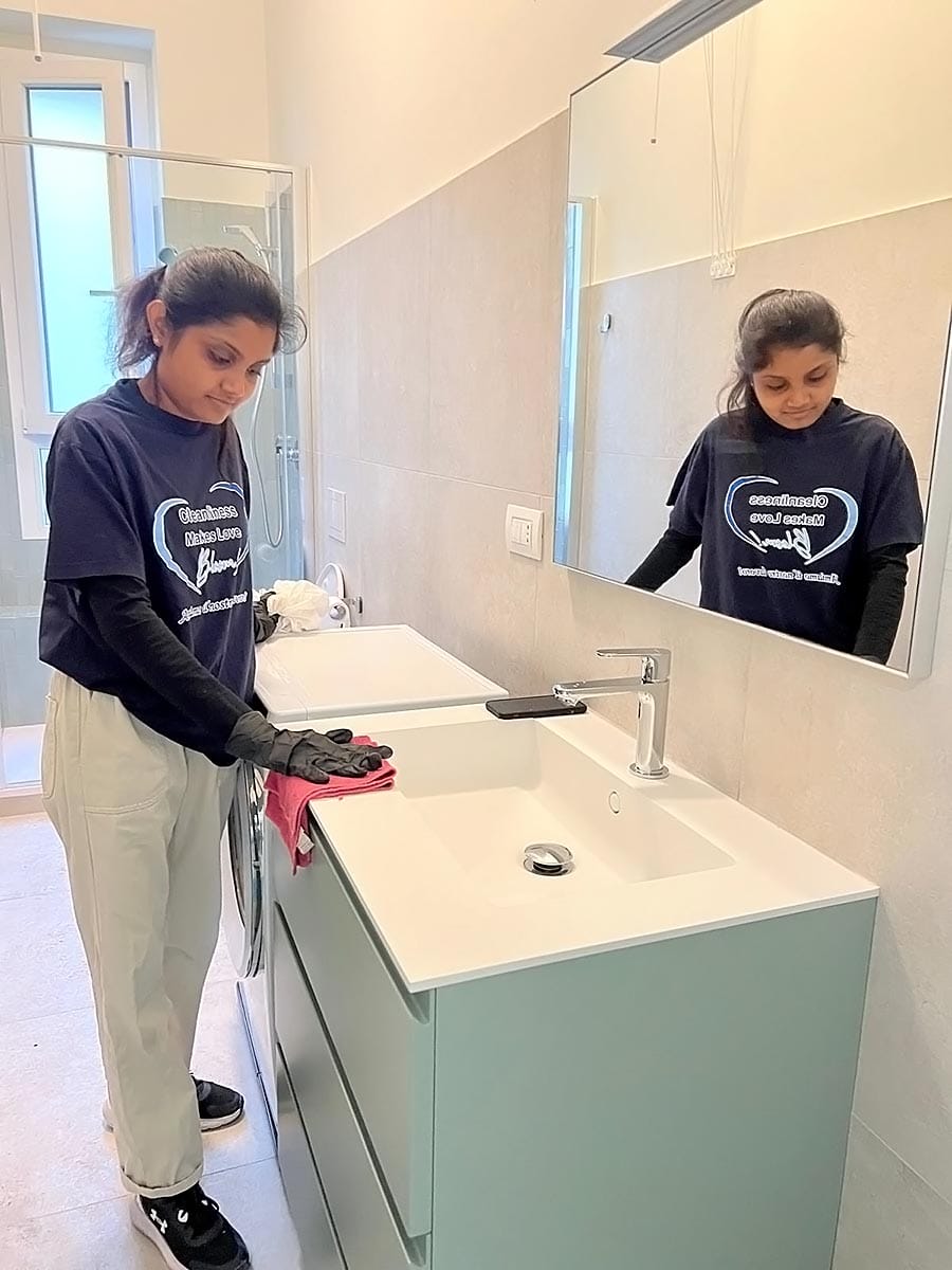 Una donna con guanti e maglietta del servizio di pulizia pulisce il lavandino del bagno con un panno rosa. Si trova in un bagno moderno con uno specchio a forma di V e una doccia in vetro sullo sfondo.