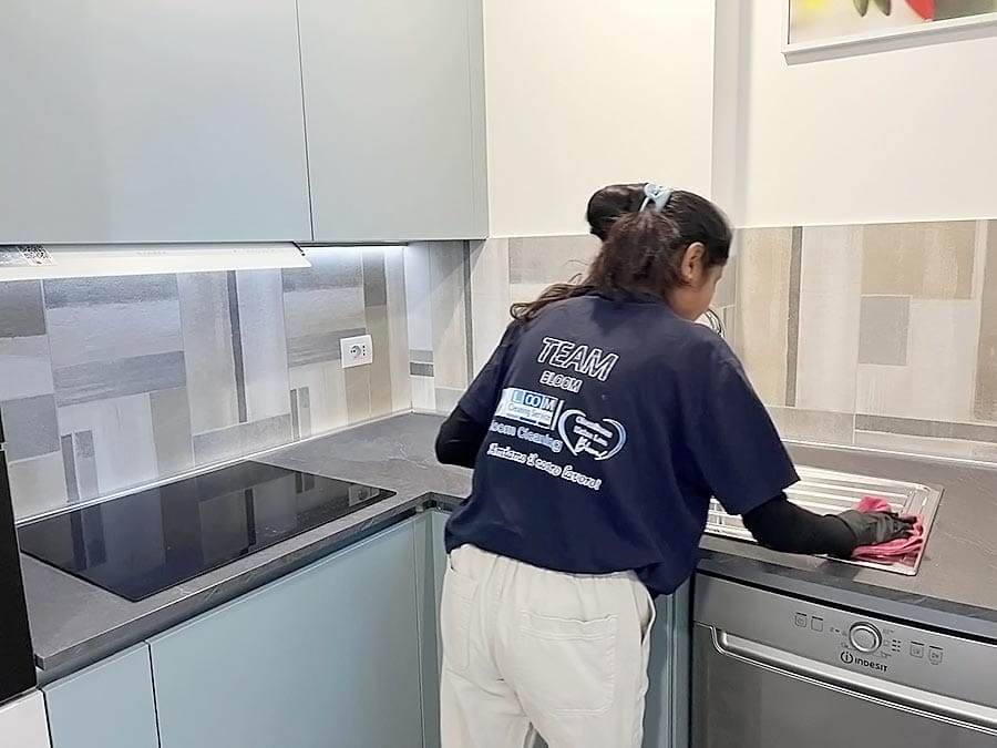 Una persona che indossa guanti e una camicia scura per le pulizie di TEAM pulisce il bancone e il lavello di una cucina moderna con armadietti azzurri, piano cottura e un'elegante cappa d'aspirazione.