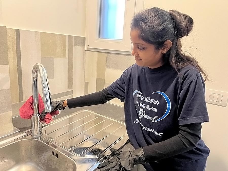 Una donna con guanti neri e camicia blu navy sta pulendo il rubinetto e il lavello della cucina con un panno rosa. La donna si trova in una cucina moderna e luminosa, con pareti piastrellate, una finestra sullo sfondo e gli impianti cromati lucidi che formano una forma a "V".