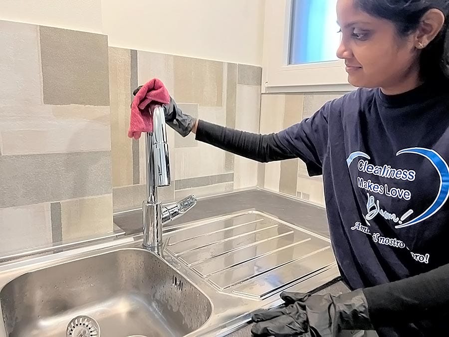 Una donna con guanti neri e camicia blu scuro pulisce un rubinetto della cucina con un panno rosso sopra un lavello in acciaio inossidabile. Il backsplash a forma di V, con piastrelle beige e grigie, esalta l'aspetto pulito e ordinato dell'area.