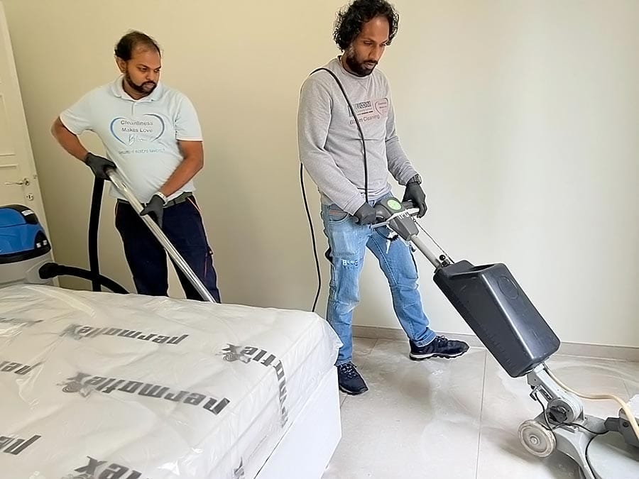 Due uomini stanno pulendo una camera da letto; uno sta usando un aspirapolvere e l'altro sta azionando una macchina per la pulizia del pavimento accanto a un letto coperto di plastica. Entrambi sono concentrati sui loro compiti nella stanza luminosa.